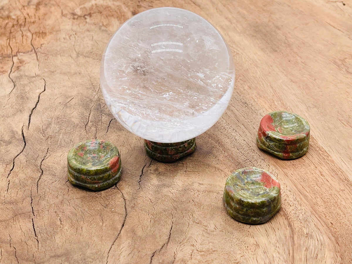 Unakite crystal sphere on carved gemstone stand, used as home decor, showcasing vibrant green and pink hues in a natural setting